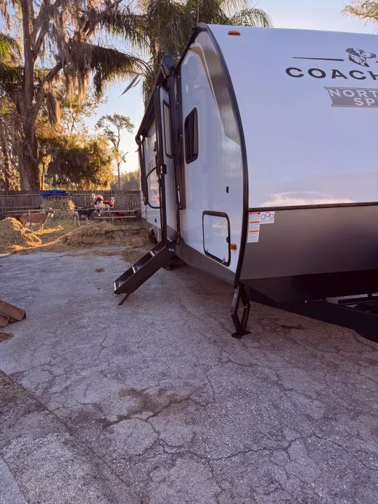 RV rental with steps down on asphalt. Goats in a hay-filled pen are visible behind it, with mossy trees in the background.. Coachmen Spirit 29BH 2026