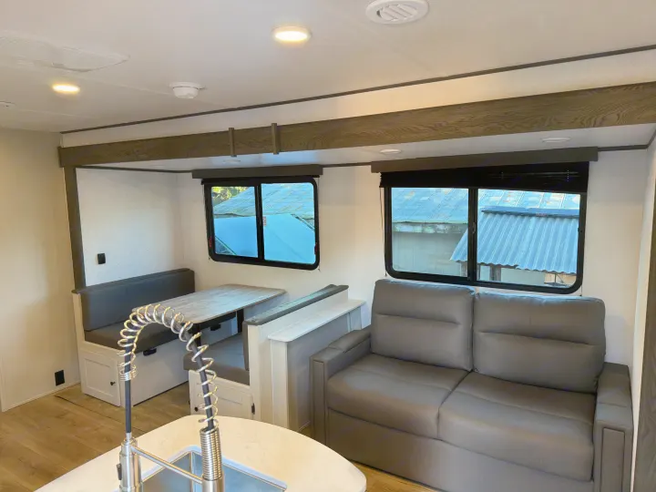 Interior of an RV rental showing a grey sofa, a light wood dinette, and a kitchen sink with a high arc faucet.. Coachmen Spirit 29BH 2026