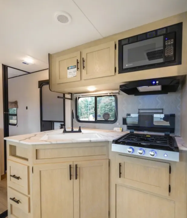 Explore this modern rv rental kitchen featuring light wood cabinets, a sink, microwave, and three-burner stovetop.. Forest River Shasta I-5 2025