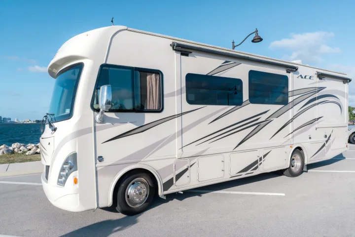 A modern, white rv rental, featuring stylish graphics, parked by a sunny waterfront with a distant city skyline.. Thor A.C.E. 2018