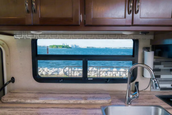 Inside an RV rental: kitchen counter, sink, and a window framing a beautiful water view with an island and city skyline.. Thor A.C.E. 2018