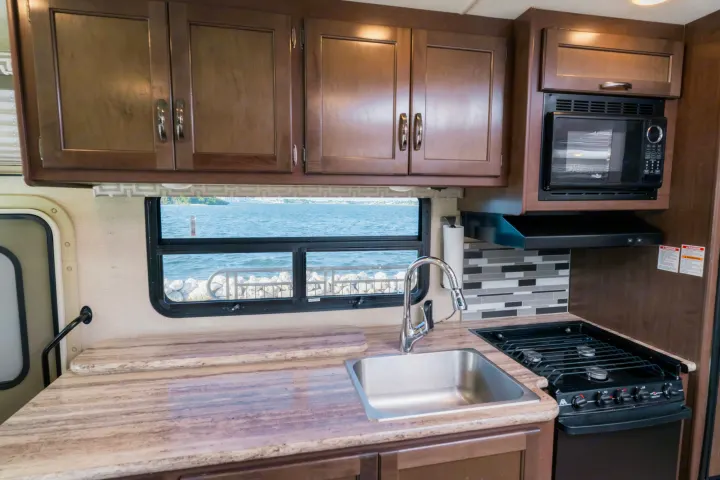 RV rental galley: microwave, stove, sink, and wood cabinets, with a tranquil lake view from the window.. Thor A.C.E. 2018