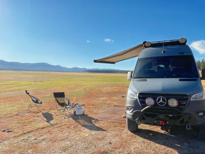 RV rental Sprinter van with awning, chairs, and fire pit in a vast, dry field with distant mountains under a blue sky.. Winnebago Revel 2022