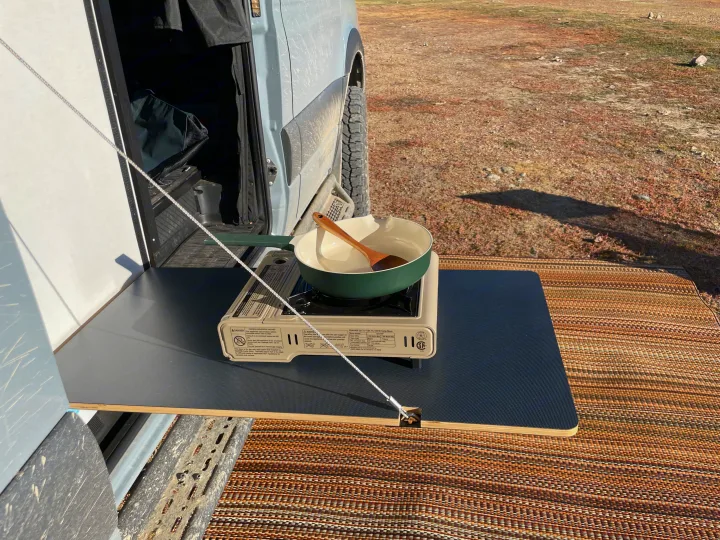 RV rental cooking setup: a portable stove, pot, and spoon on a pull-out table next to a camper van, perfect for outdoor meals.. Winnebago Revel 2022