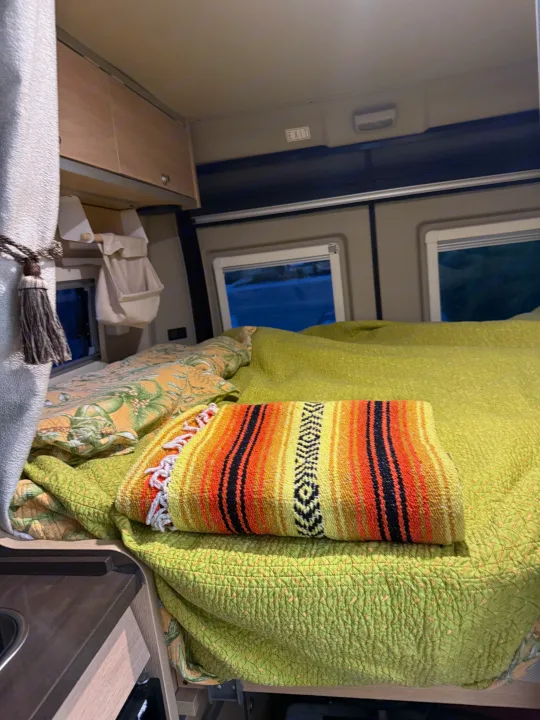 Inside a cozy **rv rental** camper van, featuring a vibrant bed with a green quilt and serape, overhead cabinets, and windows.. Hymer Hymer Class B 2019