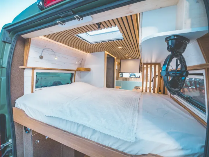 Inside a modern rv rental camper van, showing a comfy bed with white bedding, wooden walls/ceiling, and a fan.. Peugeot Boxer 2019