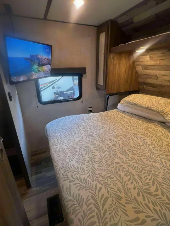 RV rental bedroom with TV displaying a coastal scene, comfy patterned bed, window view, and PS5 for cozy travels.. Crossroads RV Zinger 2024