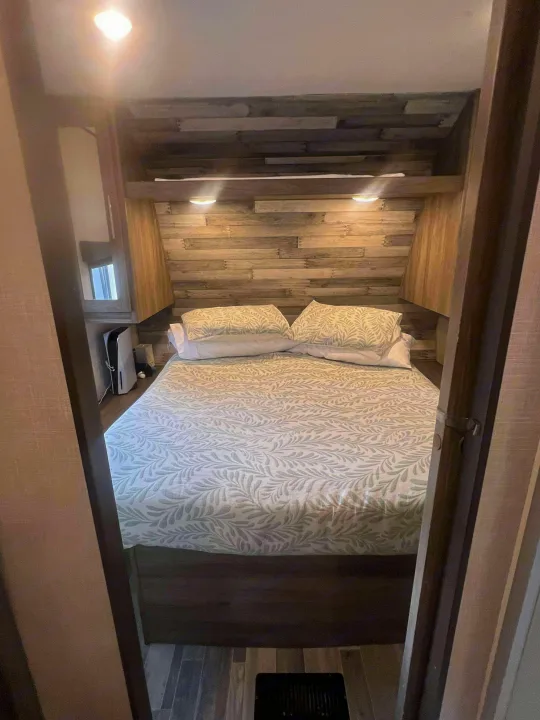 Inside this RV rental, a compact bedroom features a green leaf-patterned bed, wooden headboard with accent lights, and a PS5.. Crossroads RV Zinger 2024