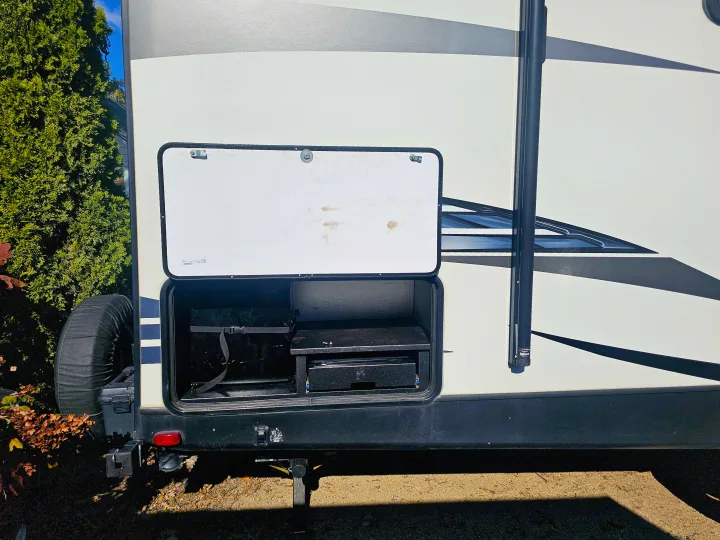 RV rental side compartment open, revealing outdoor cooking gear and a rear-mounted spare tire.. Keystone RV Outback Ultra-Lite 2021