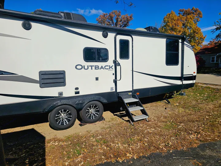 This white Outback RV rental is parked outdoors in autumn, with colorful trees and a clear blue sky overhead, ready for travel.. Keystone RV Outback Ultra-Lite 2021