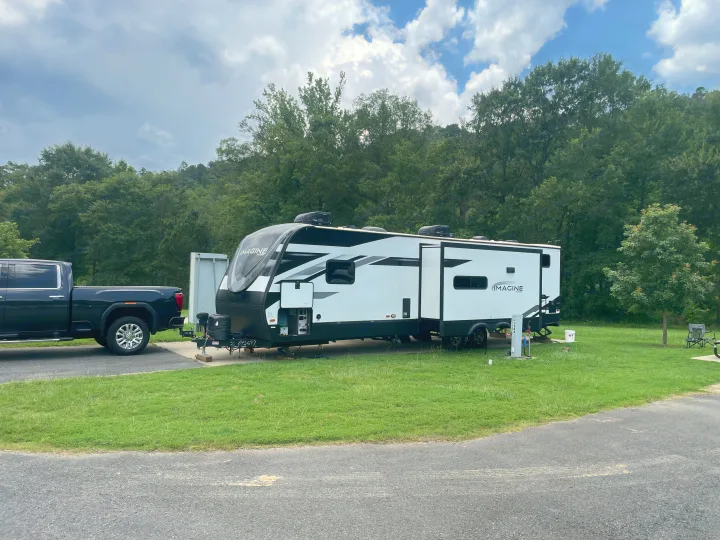 RV rental parked at a serene campground, hooked to a pickup, surrounded by green trees under cloudy skies.. Grand Design Imagine 3210BH 2023