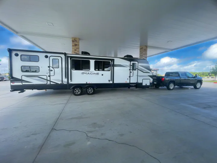 A spacious white 'IMAGINE' travel trailer towed by a black pickup truck under a gas station canopy, an ideal rv rental setup.. Grand Design Imagine 3210BH 2023