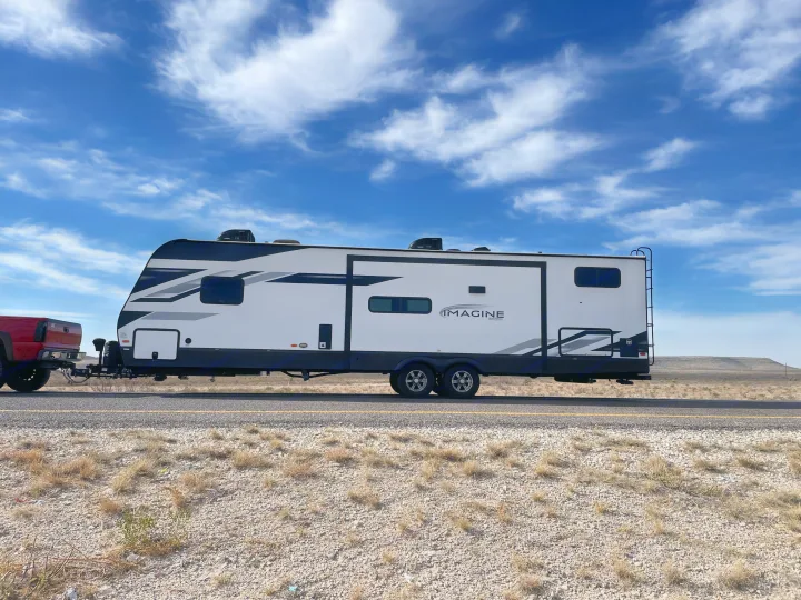 A white Grand Design Imagine travel trailer RV rental is being towed by a red truck on a highway under a blue sky.. Grand Design Imagine 3210BH 2023