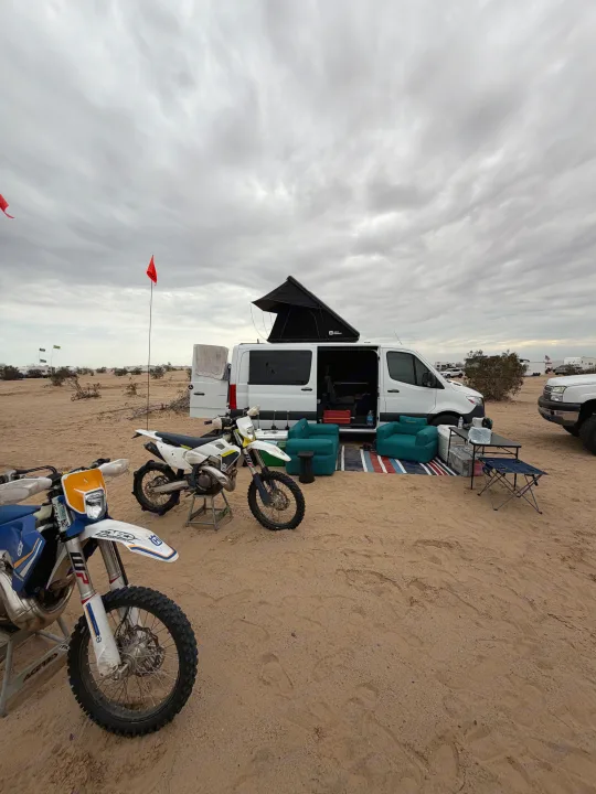 Adventure-ready RV rental with dirt bikes parked in a sandy desert campsite under an overcast sky, ideal for off-road fun.. Mercedes Sprinter 2024