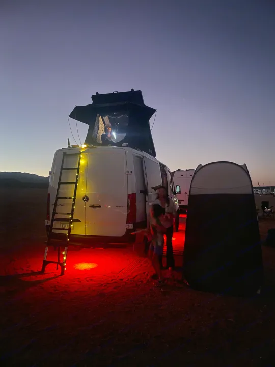 Dusk adventure with an RV rental van. Illuminated rooftop tent, ladder lights, and red ground lights. People & privacy tent for van life camping.. Mercedes Sprinter 2024
