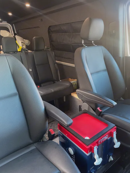 Inside this RV rental, multiple rows of black seats, a red and blue RTIC cooler, and upholstered walls are visible.. Mercedes Sprinter 2024