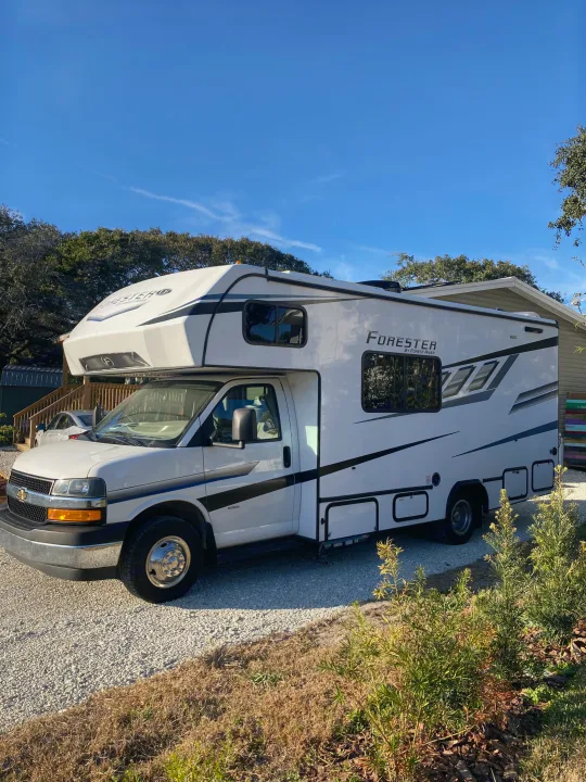Bright blue sky above a white Forester Class C RV rental parked on a gravel driveway, ready for your next travel adventure.. Forester LE 2024