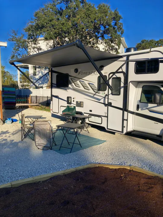 RV rental parked on gravel, with its awning extended over a comfy outdoor setup including a portable grill and folding chairs.. Forester LE 2024