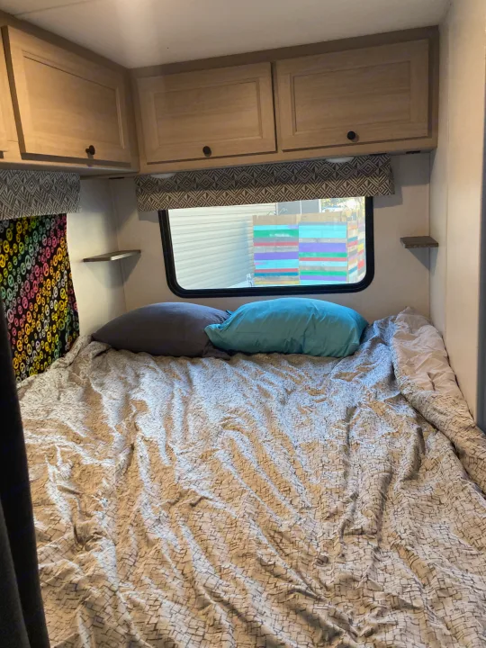 An RV rental bedroom features a bed with grey and blue pillows, patterned bedding, overhead cabinets, and a window.. Forester LE 2024