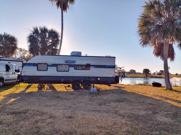 A "Salem Cruise Lite" RV rental by a lake, featuring palm trees, a dog, and bikes, ready for a sunny getaway.. Forest River Salem Cruise Lite 2021