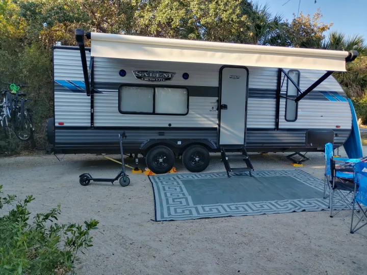 A spacious RV rental is parked with an awning, outdoor rug, chairs, electric scooter, and bikes at a sunny campsite.. Forest River Salem Cruise Lite 2021