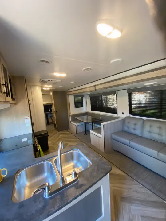 Bright RV rental interior showcasing a modern kitchen with a double sink, a U-shaped dining booth, and a gray sofa.. Keystone RV Cougar 2022