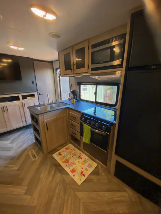 This cozy RV rental kitchen features light wood cabinets, a double sink, stove, microwave, and fridge on a herringbone floor.. Keystone RV Cougar 2022