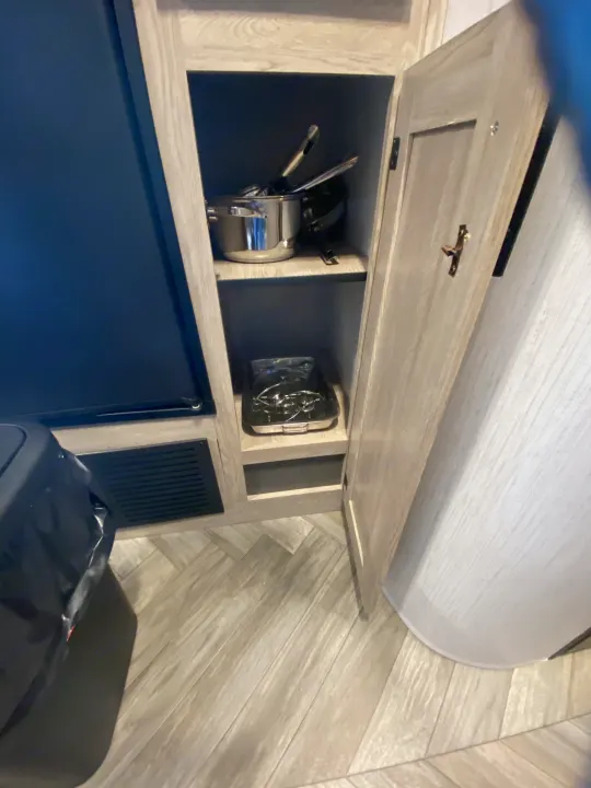 Inside an RV rental, a light-wood cabinet reveals kitchen essentials: pots, pans, and a portable stovetop. The floor has a herringbone pattern.. Keystone RV Cougar 2022