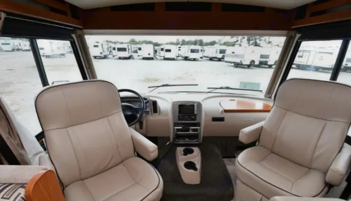 Inside a bright RV rental. View from the cockpit shows light-colored seats, a modern dashboard, and many other RVs parked in a lot ahead.. Winnebago Vista 2019