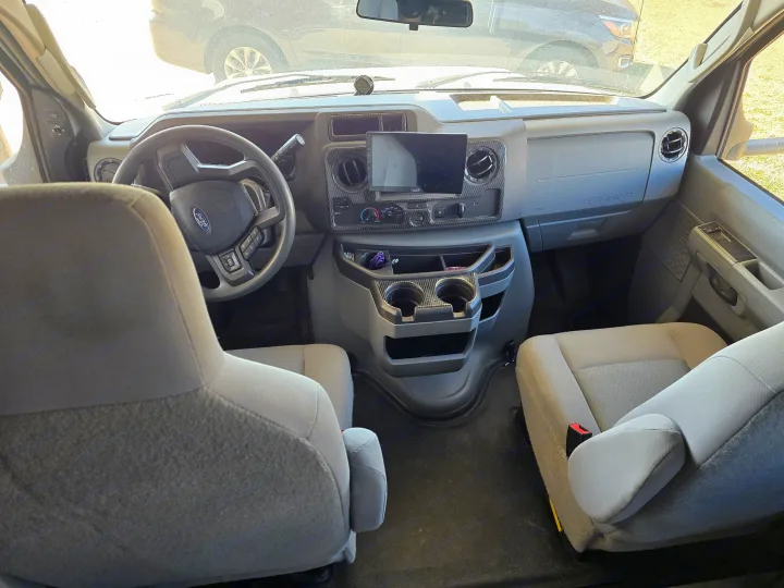 Interior of an RV rental van's front cabin, showing the Ford steering wheel, dashboard with infotainment screen, and gray seats.. Forest River Coachmen Leprechaun Premier 2022