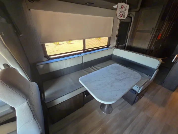 Bright RV rental dinette features two-tone seating, a marble-top table, and a large window with shades pulled.. Forest River Coachmen Leprechaun Premier 2022