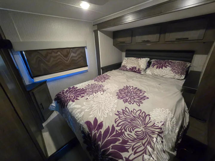 RV rental bedroom, offering a cozy bed with purple floral bedding, convenient overhead cabinets, and ambient window lighting.. Forest River Coachmen Leprechaun Premier 2022