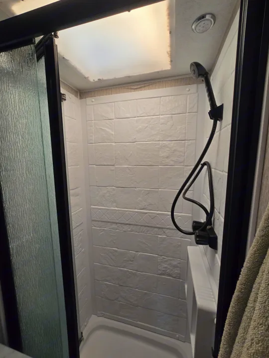 Compact RV rental shower with white textured walls, handheld showerhead, bright skylight, and frosted glass door.. Forest River Coachmen Leprechaun Premier 2022