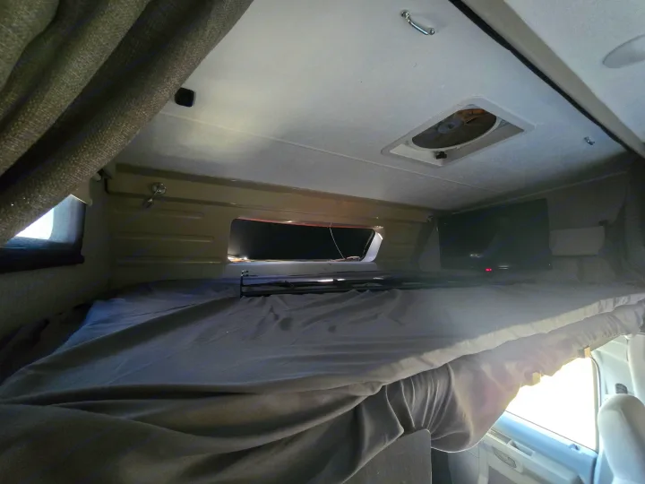 Inside an rv rental, showing the cozy overcab bunk bed with grey sheets, a TV, ceiling vent, and window.. Forest River Coachmen Leprechaun Premier 2022