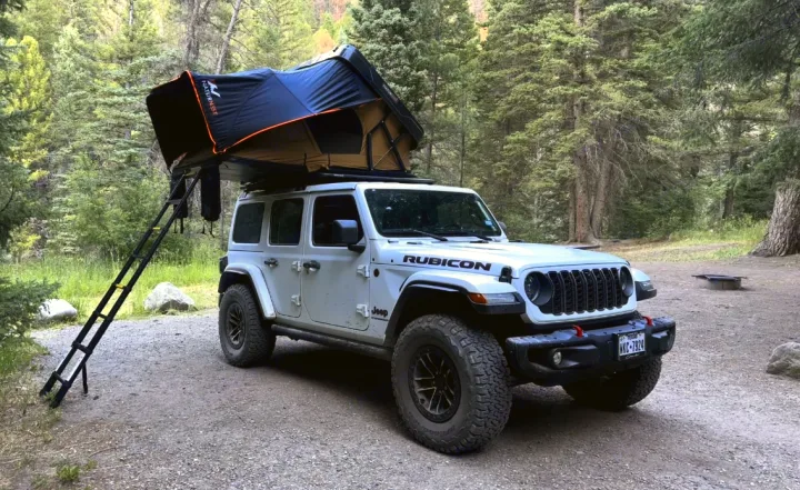 White Jeep Wrangler with a rooftop tent set up in a forest. Experience outdoor camping with this unique RV rental.. Jeep Wrangler Rubicon X 2024
