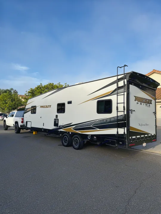 A large white Powerlite Prime toy hauler, perfect for an rv rental, hitched to a white pickup truck on a residential street.. Genesis Supreme Powerlite LE Toy Hauler 2022
