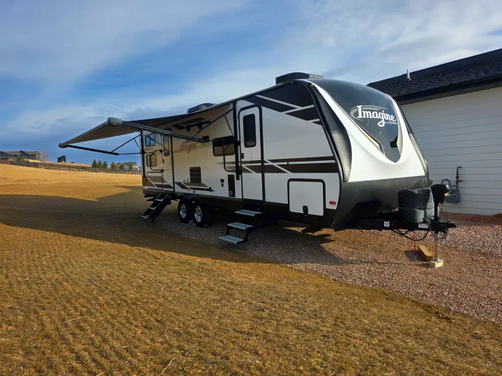 White & black Imagine travel trailer, perfect for your next RV rental adventure. Awning extended, parked on gravel in a sunny field.. Grand Design Imagine 2022
