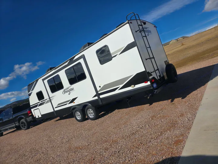 RV rental: A white Imagine travel trailer, hitched to a black pickup, is parked on a gravel lot under a bright blue sky.. Grand Design Imagine 2022