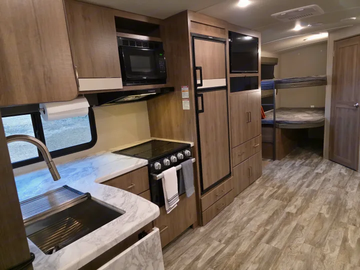 Modern rv rental interior: kitchen with stove, microwave, fridge, sink, and bunk beds, featuring wood-grain finishes.. Grand Design Imagine 2022