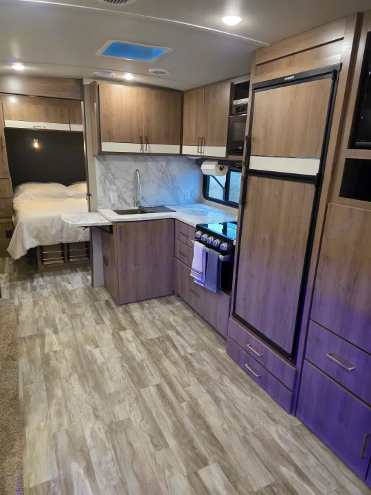 Modern rv rental interior showcasing a compact kitchen with wood-grain cabinets, sink, stove, integrated fridge, and cozy bed area.. Grand Design Imagine 2022