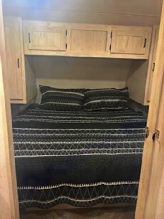RV rental bedroom features a comfy queen bed with dark geometric bedding, flanked by light wood cabinets and overhead storage.. East to West Della Terra 2023