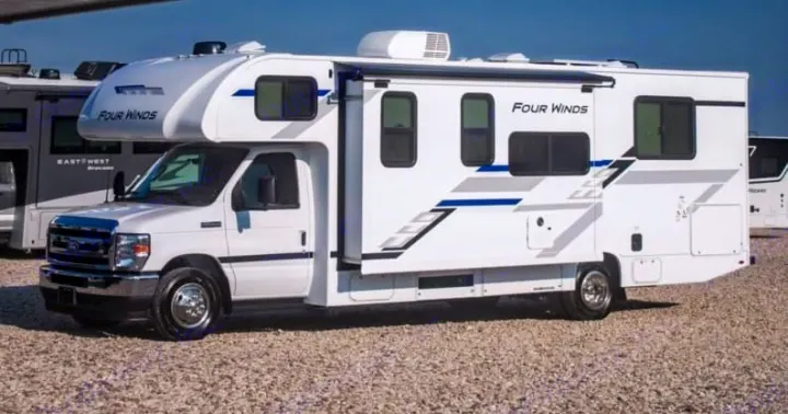 A white Four Winds Class C RV, with blue and grey decals and an extended slide-out, parked on gravel. Ideal rv rental.. Thor four winds 2027