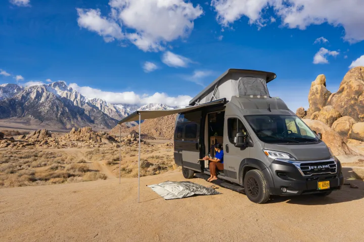 Experience van life! An RV rental camper van is parked in a scenic mountain desert with snow-capped peaks. A woman relaxes by the open door under an awning.. Coachmen PIXEL 2026
