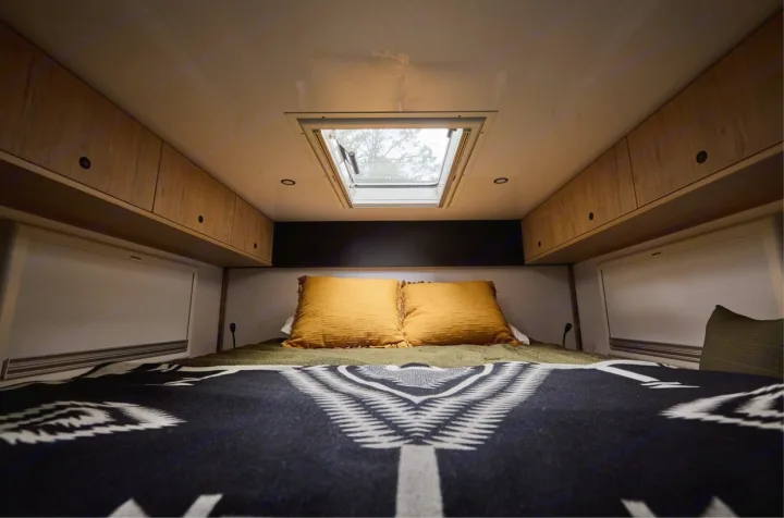 Cozy RV rental bedroom: bed with yellow pillows, patterned blanket, skylight, and wood overhead cabinets.. Storyteller Epic 2025
