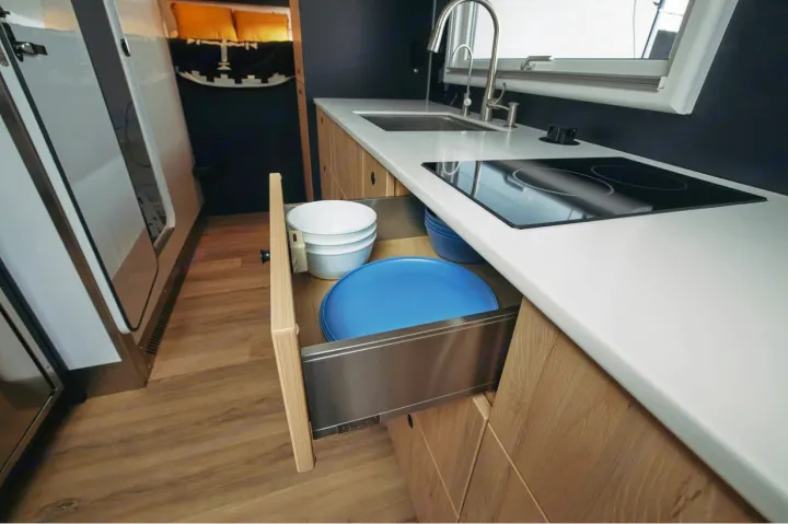 Modern RV rental kitchen. Features a white countertop, sink, induction hob, and an open drawer with dishes inside.. Storyteller Epic 2025
