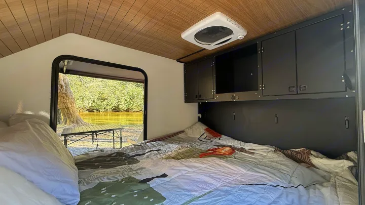 Cozy RV rental interior with bed, overhead storage, and a scenic view of a river/lake and trees outside the open door.. Cedar Ridge Deneb XT 2025