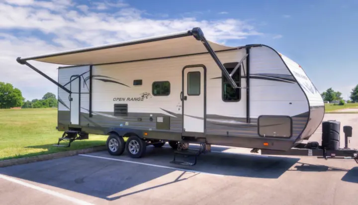 White & gray Open Range travel trailer RV rental with awning extended, parked on asphalt by a grassy field on a sunny day.. Highland Ridge RV Open Range 2019