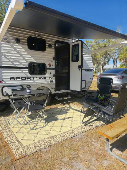 An RV rental with its awning extended creates a cozy outdoor living space with a rug, table, chairs, and a Tesla parked nearby.. KZ Sportsmen Classic 130BH 2026