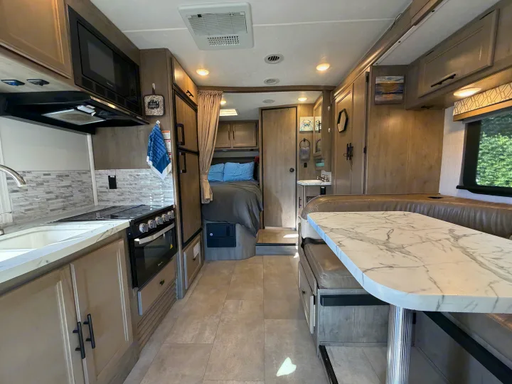Modern rv rental interior: kitchen with stove/oven, dining booth with marble-look table, and a cozy bedroom. Light-toned wood and flooring.. Coachmen Other 2020
