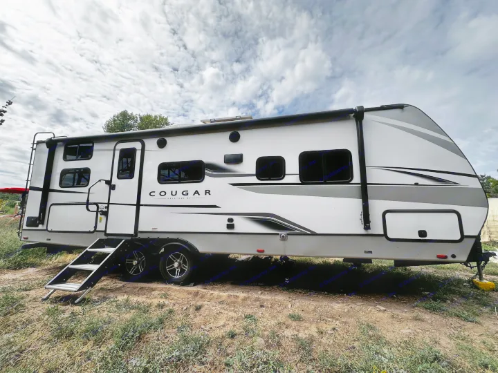 This white Cougar travel trailer RV rental is set outdoors, featuring steps, dark windows, and a rear ladder.. Keystone RV Cougar 2023